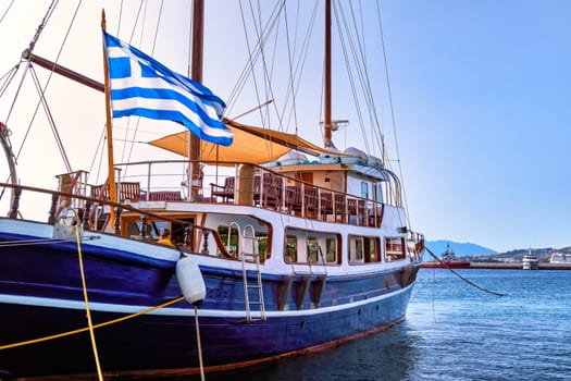 Schooner or sailboat under Greek flag moored at jetty of typical Greek island. Beautiful whitewashed houses on hills and by waterfront of harbor. Summer day sunshine, vacations and adventures. Mykonos, Greece.
