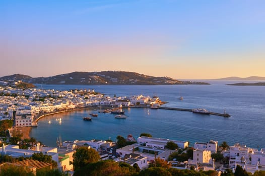 Beautiful sunset view over Chora, main town of Mykonos, Cyclades, Greece and its harbor and port. Golden hour, harbor, cruises, ship, whitewashed houses and distant islands in hazy background. Vacations. leisure, Mediterranean lifestyle