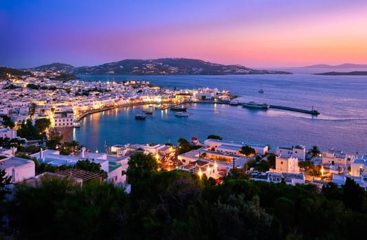 Iconic view after sunset over Chora, main town of Mykonos, Cyclades, Greece and its horseshoe-shaped harbor and port. Harbor, cruises, ship, whitewashed houses and distant islands in hazy background. Town lights up. Vacations, Mediterranean lifestyle