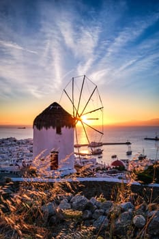 Famous traditional white windmill overlook civil port and harbor of Mykonos, Cyclades, Greece at sunset sky. Beautiful sky, sun touch sea horizon, Chora, main town of island, in evening lights and shadows. Vertical shot
