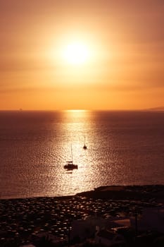 Superb sunset seascape. Low sun in hazy sky and sun glare on calm sea waters. Silhouettes of small sailboats go to sea in sunlight strip, dark coast in foreground. Summer hot days, vacations, leisure, romantic sailing. Vertical shot