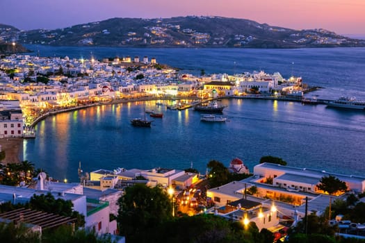 Iconic view after sunset over Chora, main town of Mykonos, Cyclades, Greece, horseshoe-shaped harbor and port. Bay, cruises, ship, whitewashed houses and distant islands in hazy background. Town lights up. Vacations, leisure, Mediterranean lifestyle