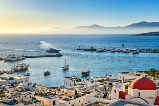 Beautiful view of Chora, Mykonos, Greece at sunset. Civil port in harbor, boats, sailboats, yachts moored by jetty. Famous whitewashed houses and traditional white church with red dome overlooking the hazy horizon and islands