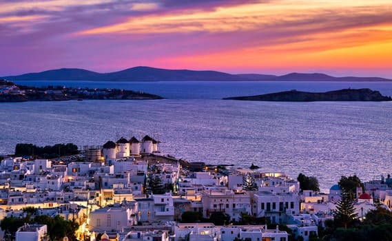 Beautiful sunset view of famous traditional white windmills on hilltop, Mykonos, Greece. Whitewashed houses, distant islands. Colorful sunset sky, hazy summer evening, iconic destination, town lights on. Vacations, leisure, Mediterranean lifestyle