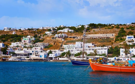 Beautiful summer day and sunshine in typical marina of Greek island. Colorful fishing boats and tourist cruise yachts moored at jetty. Whitewashed houses on hills and by waterfront. Mediterranean lifestyle, vacations. Mykonos, Cyclades, Greece.
