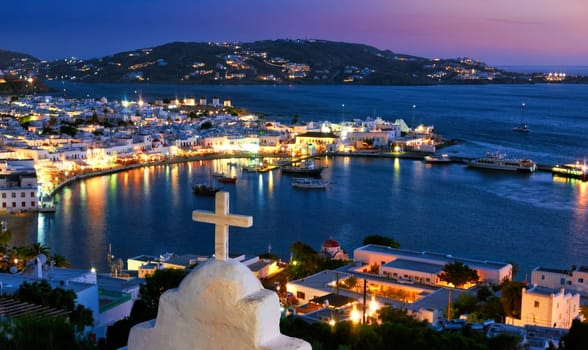 Beautiful view over Chora, main town of Mykonos, Cyclades, Greece after sunset from rooftop of Greek Orthodox church and its white marble cross, bay, ships, whitewashed houses. Town lights up. Vacations. leisure, Mediterranean lifestyle
