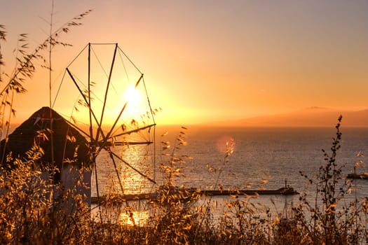 Golden sunset over sea horizon in Mykonos, Cyclades, Greece. Famous traditional white windmill overlook civil port and harbor on hot summer evening in bright sunlight. Romantic image, iconic travel destinations