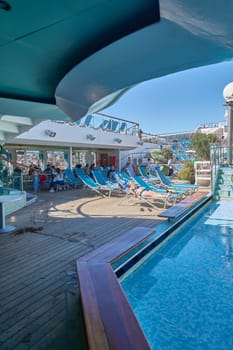 Catania, Italy - February 18, 2025: A woman sunbathes on a deck chair near a swimming pool on a cruise ship Other passengers enjoy drinks and conversations under the sun. Concept: vacation, relaxation