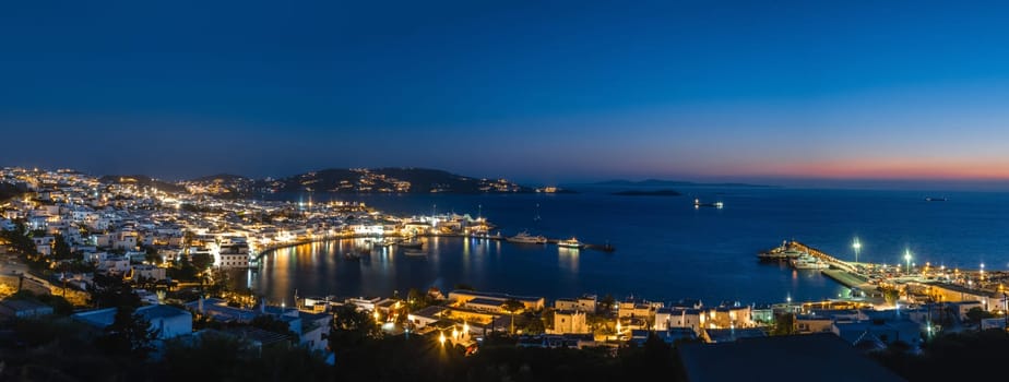 Iconic night panorama over Chora, main town of Mykonos, Cyclades, Greece. Harbor, ships, cruises, whitewashed houses and distant islands in hazy background. Town lights up. Vacations, leisure, nightlife, Mediterranean lifestyle