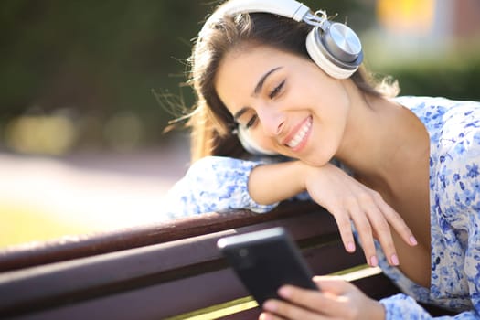 Happy female listening to music with phone and headphone sitting in a bench in a park