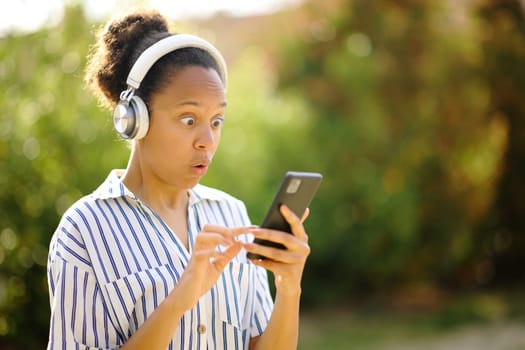 Surprised black woman wearing headphone listening to music on phone in a park