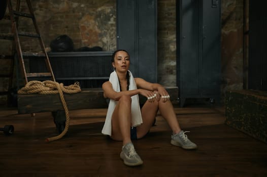 Woman boxer resting on the floor with towel on neck. Young tired girl in sportswear sitting and looking at camera over old gym grungy interior