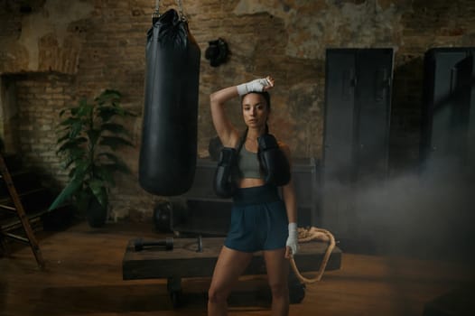 Serious woman boxer posing for camera dressed in sportswear, hands bandages carrying boxing gloves on shoulders standing over loft gym interior