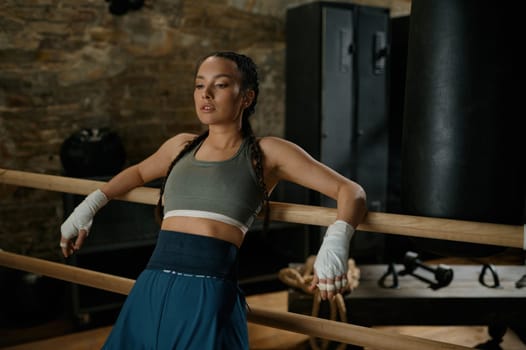 Athletic female fighter with bandaged hands standing leaned at wooden training bar in the loft gym interior