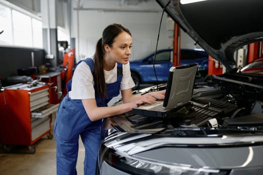 Young woman car engineer providing computer diagnostics using laptop with special interface