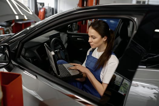 Young woman technician sitting in car cabin working on laptop providing computer diagnostics. Auto service station