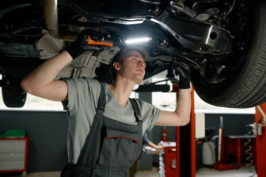 Male auto mechanic working under car lifted on elevator with flashlight at garage service