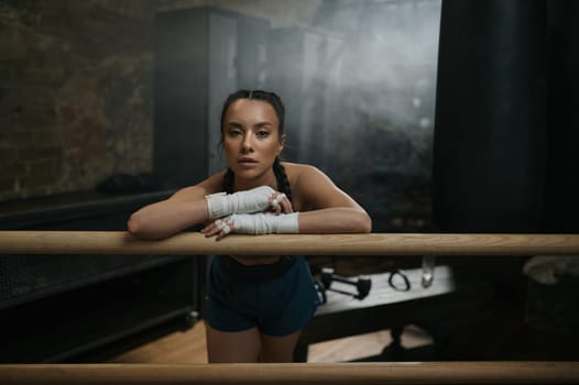 Pretty young woman boxer resting at boxing ring ready for workout. Tired female fitness athlete exercising and relaxing before or after strong sports training