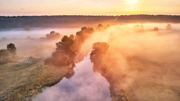 Summer foggy sunrise landscape panorama. River flowing in rural field, surrounded by lush green trees. Water reflecting warm orange glow of rising sun. Yellow misty light