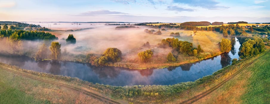 Aerial summer sunrise view. Rural landscape, river meandering in meadow lush green trees. Morning Foggy Scene. Serene peaceful atmosphere. Small farmland village in distance. Beauty of nature, tranquility of countryside.