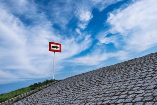 Black arrow on white background and in red frame standing atop of cobblestone paved hill at day. Blue sky with great clouds. Copy space, concept of direction, choice, signal, symbol, hope, options, opportunities, right