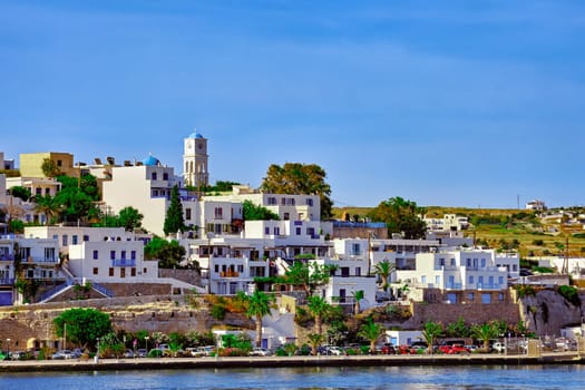 Beautiful summer day and sunshine in typical town of Greek island. Whitewashed houses on hills and by waterfront. Orthodox church. Mediterranean lifestyle, vacations. Milos, Cyclades, Greece.