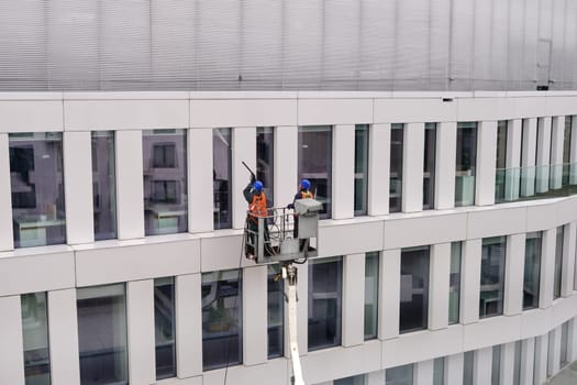 Two workers wearing safety harness wash walls, panels and windows of office building facade at height standing in a crane cradle or aerial platform using pressure washer, wipers and mops. Daytime, Europe, cleaning facades, modern architecture.