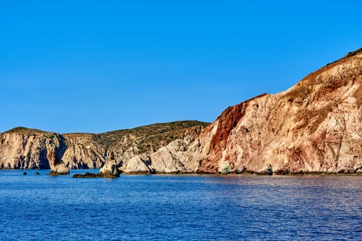 Beautiful view of high hill slope of island in Mediterranean Sea. Blue sea waters, clear blue sky, bright sunshine, summer day, rocks and islets of Aegean sea.