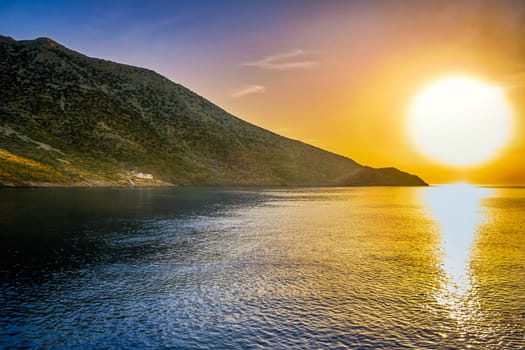 Beautiful view of Mediterranean island and its coast at sunset. Big low sun, sun flares and sun rays beam on lonely white Orthodox church on seashore. Milos island, Cyclades, Greece