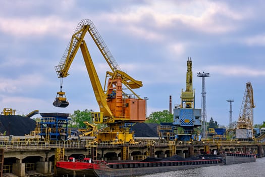 Close view of river port cranes with clamshell or griper loading coal to river drag boats or barges moored by pier on cloudy day. Problems of energy production, natural mineral deposits, environment crisis, alternatives