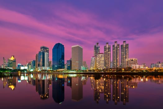Beautiful landscape with modern high-rise buildings reflecting in calm lake waters at colorful sunset. Benjakitti Park, Bangkok, Thailand.
