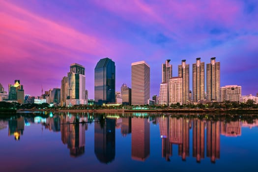 Beautiful landscape with modern high-rise buildings reflecting in calm lake waters by park and promenade at colorful sunset. Benjakitti Park, Bangkok, Thailand.