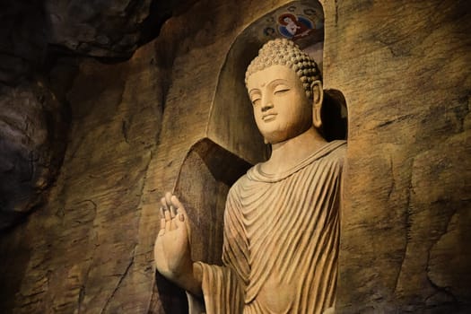 Statue of standing Buddha in monk clothes carved in limestone cave and lit by light. Right hand in adhay mudra gesture. Religion, tradition, prayer, meditation.