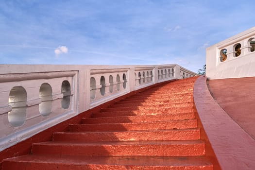Red sunlit stairs and white banisters leading upwards at blue sky. Concept of ascension, opportunity, richness. Steps up to temple of Wat Saket or Golden Mount, Bangkok, Thailand.