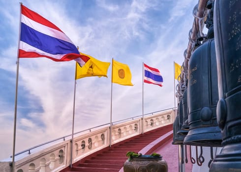 Row of Thai national, buddhism and Thai Royal flags along spiral staircase of red steps and white parapet, bells in blurred foreground. Stairway at Golden Mount temple or Wat Saket, Bangkok, Thailand. Incomplete inscriptions in Thai not translable.