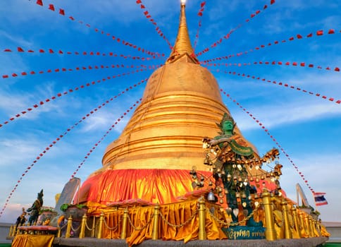 Statue of green-skinned Thai angel holding roll with message in Thai - translation: free from sorrow - stand in front of big golden stupa in sunlight and at blue sky with clouds in Buddhist temple. Decorations with bells, golden leaves and red flags.