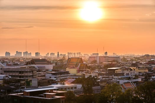Beautiful cityscape of Bangkok city center in low sun, hazy orange sky and clouds, Thailand. Residential low-rise area in foreground, traditional temples, high-rises in downtown. Heat, summer, haze, heatwave, Asian climate, Buddhist religion.