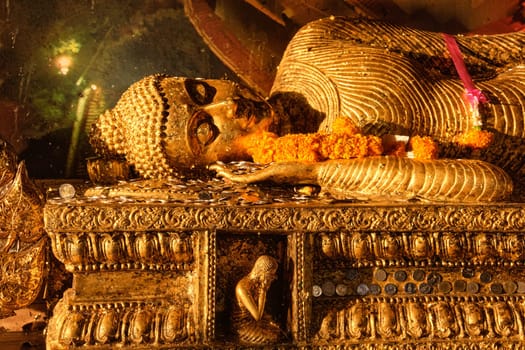 Statue of reclining Buddha covered with golden leaves, coins and garlands of flowers as offerings in soft sunlight. Traditional image, rituals of prayer, worship and blessing. Buddhism, religion, Theravada, Thailand, sacred.