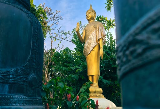 Gilded statue of standing Buddha in monk clothes and covered by golden textile shot between two bells and among trees and flowers. Right hand in adhaya mudra gesture. Selective focus, blurred foreground, religion, tradition, prayer, meditation.