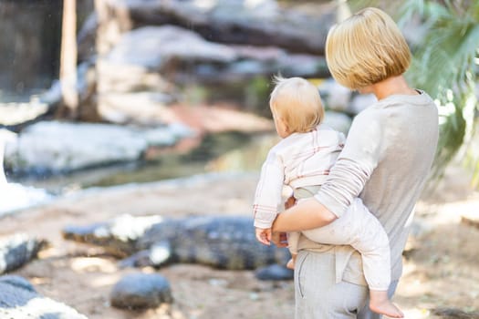 Caucasian blonde mother holding her infant baby boy child in her lap watching crocodile in zoo. Happy family having fun with animals safari park on warm summer day.