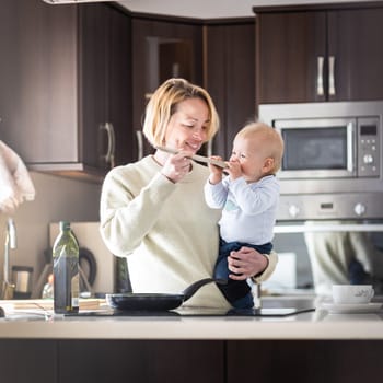 Happy mother and little infant baby boy cooking and tasting healthy dinner in domestic kitchen. Family, lifestyle, domestic life, food, healthy eating and people concept