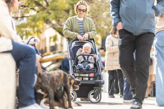 Mother walking and pushing his infant baby boy child in stroller in crowd of people wisiting sunday flea market in Malaga, Spain