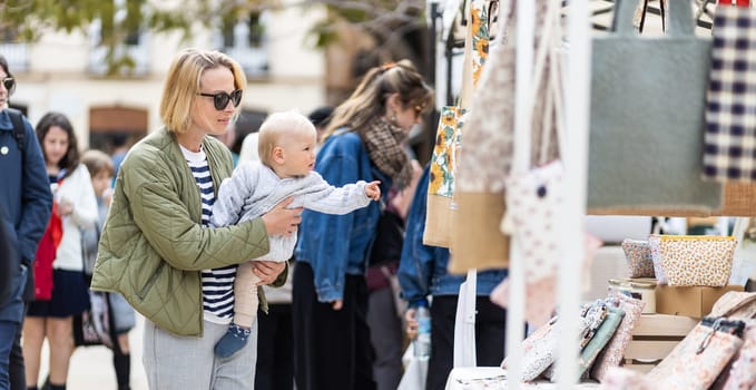 Mother walking carrying his infant baby boy child in crowd of people wisiting sunday flea market in Malaga, Spain.
