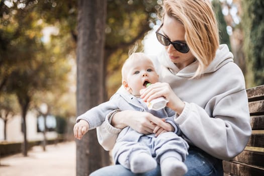 Mother sitting on bench in urban park, holding her infant baby boy child in her lap and feeding him baby food on nice spring sunny day.
