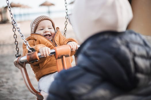 Mother pushing her cheerful infant baby boy child on a swing on sandy beach playground outdoors on nice sunny cold winter day in Malaga, Spain