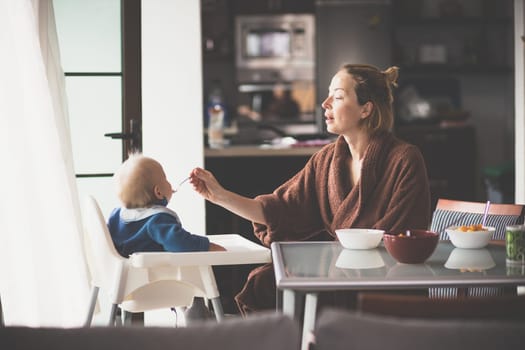 Mother wearing bathrope spoon feeding her infant baby boy child sitting in high chair at the dining table in kitchen at home in the morning.
