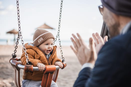 Father pushing hir cheerful infant baby boy child on a swing on sandy beach playground outdoors on nice sunny cold winter day in Malaga, Spain