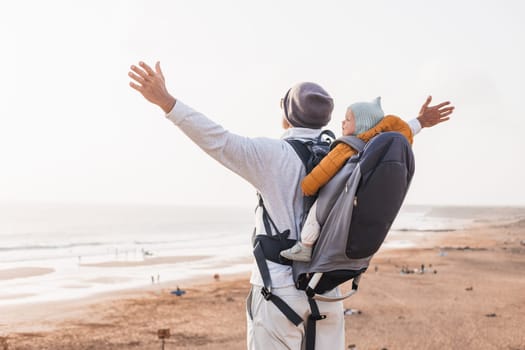 Young father rising hands to the sky while enjoying pure nature carrying his infant baby boy son in backpack on windy sandy beach. Family travel concept