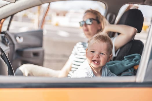 Mother and her infant baby boy child on family summer travel road trip, sitting at dad's front seat, waiting in the car for father to buy farry tickets