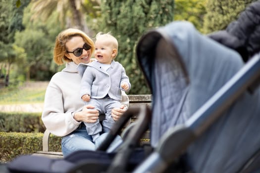 Mother sitting on bench in urban park, laughing cheerfully, holding her smiling infant baby boy child in her lap having baby stroller parked by their site.